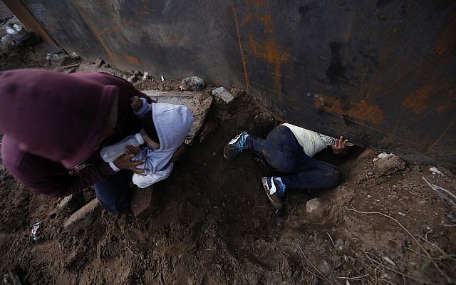 El migrante hondureño Joel Méndez, de 22 años, se fue, alimenta a su hijo Daniel de ocho meses de edad, mientras que su compañera Yesenia Martínez, de 24 años, se arrastra a través de un agujero debajo del muro fronterizo de los EE. UU., En Tijuana, México, el 7 de diciembre de 2018. (Rebecca Blackwell / AP)