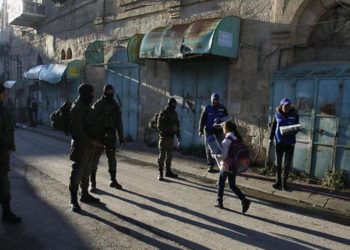 Observadores palestinos, a la derecha, observan cómo los niños pasan junto a los soldados israelíes en su camino a la escuela en la ciudad cisjordana de Hebrón