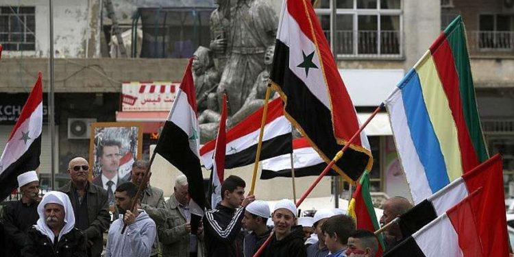 Los residentes de los Altos del Golán ondean banderas sirias y drusas mientras se reúnen frente a un retrato del presidente sirio Bashar Assad durante una protesta contra el reconocimiento del presidente estadounidense Donald Trump de la soberanía israelí sobre el área en disputa, en la ciudad de Majdal Shams, el 23 de marzo. , 2019. (Jalaa Marey / AFP)