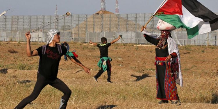 Un terrorista lanza piedras a las tropas israelíes mientras una mujer vestida con un traje tradicional palestino ondea una bandera nacional, durante los ataques cerca de la frontera con Israel, al este de la ciudad de Gaza, el 10 de mayo de 2019. (Dijo KHATIB / AFP)