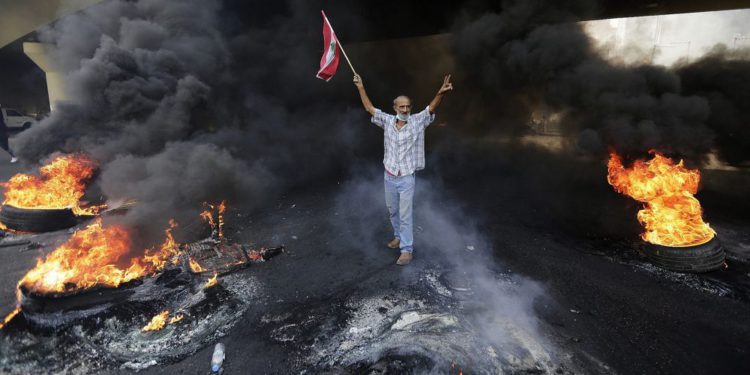 Un manifestante antigubernamental hace la señal de la victoria, mientras sostiene una bandera nacional libanesa y camina quemando neumáticos bloqueando una carretera durante una protesta contra los planes del gobierno de imponer nuevos impuestos en Beirut, Líbano, 18 de octubre de 2019. (Foto AP / Hassan Ammar )