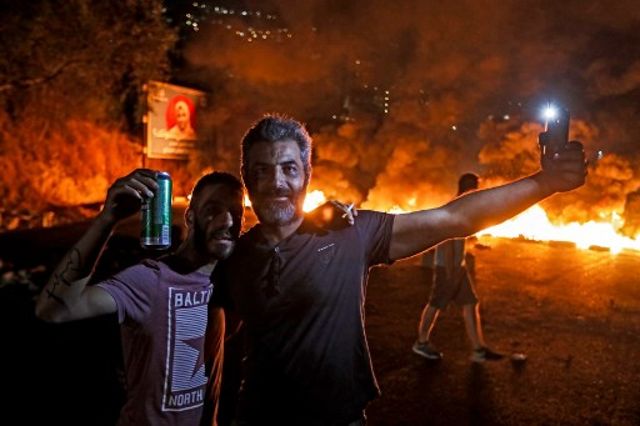 Un hombre sostiene su teléfono celular con el destello que ilumina su rostro mientras otro junto a él levanta una lata de cerveza mientras está parado frente a un incendio de llantas que bloquea la carretera principal Beirut-Trípoli, cerca de la ciudad de Tabarja, al norte de la capital, en medio de protestas contra terribles condiciones económicas el 18 de octubre de 2019. (Joseph Eid / AFP)