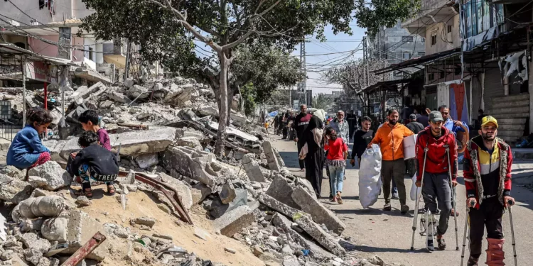 Hombres heridos caminan con muletas junto a niños sentados sobre los escombros de un edificio derrumbado en una calle de Rafah, en el sur de la Franja de Gaza, el 9 de abril de 2024. (AFP)