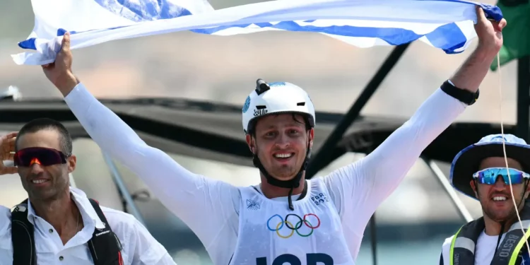 El israelí Tom Reuveny celebra con la bandera nacional después de ganar la medalla de oro en la final masculina de windsurf IQFoil en la competición de vela de los Juegos Olímpicos de París 2024, en la Marina Roucas-Blanc en Marsella el 3 de agosto de 2024. (Christophe SIMON / AFP)